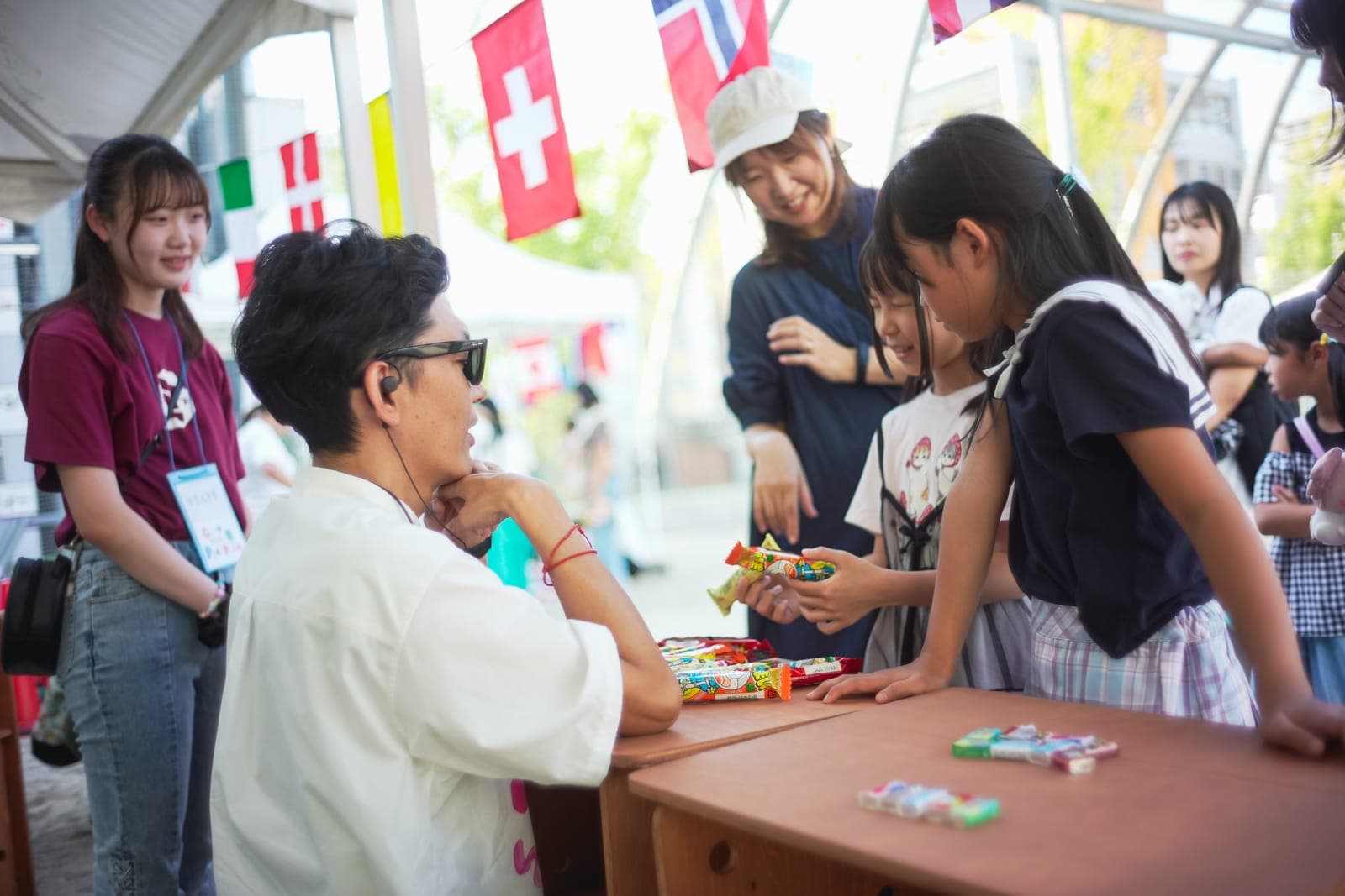 国旗の飾られたテントの下で、サングラスをかけた店主が駄菓子の並ぶ台の前に立ち、子どもたちが手探りでお菓子を選んでいる。